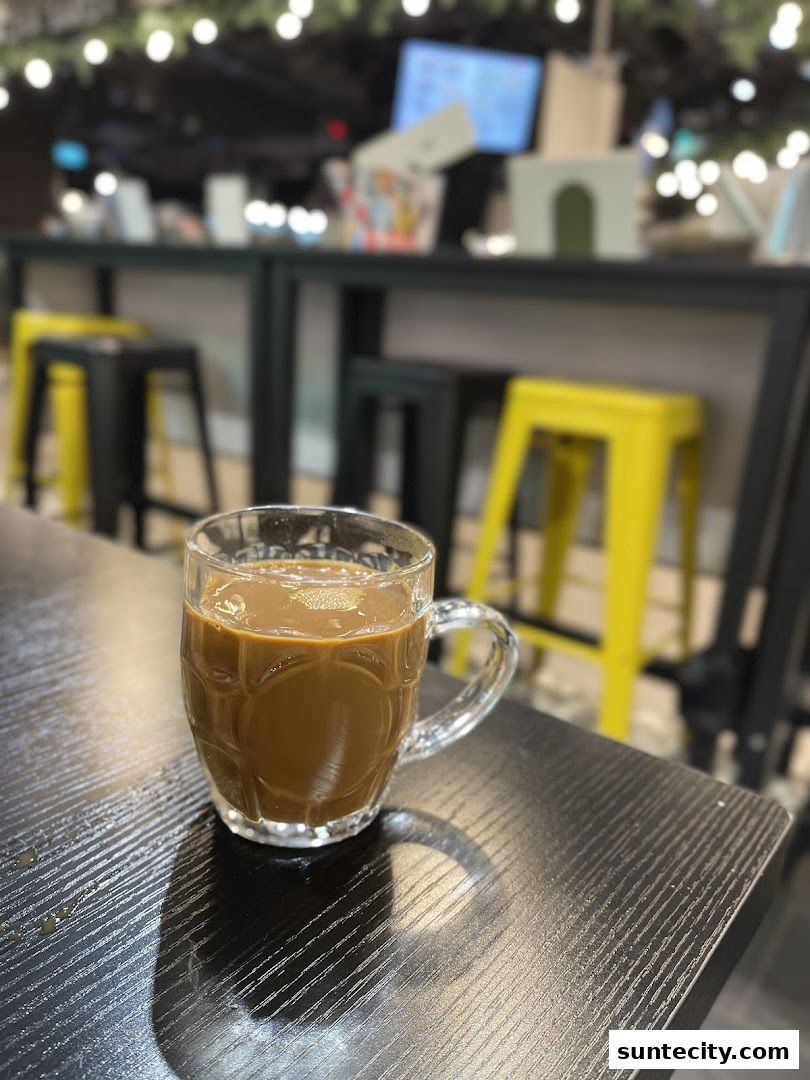 A glass mug of coffee sits on a dark wooden table with yellow stools in the background.