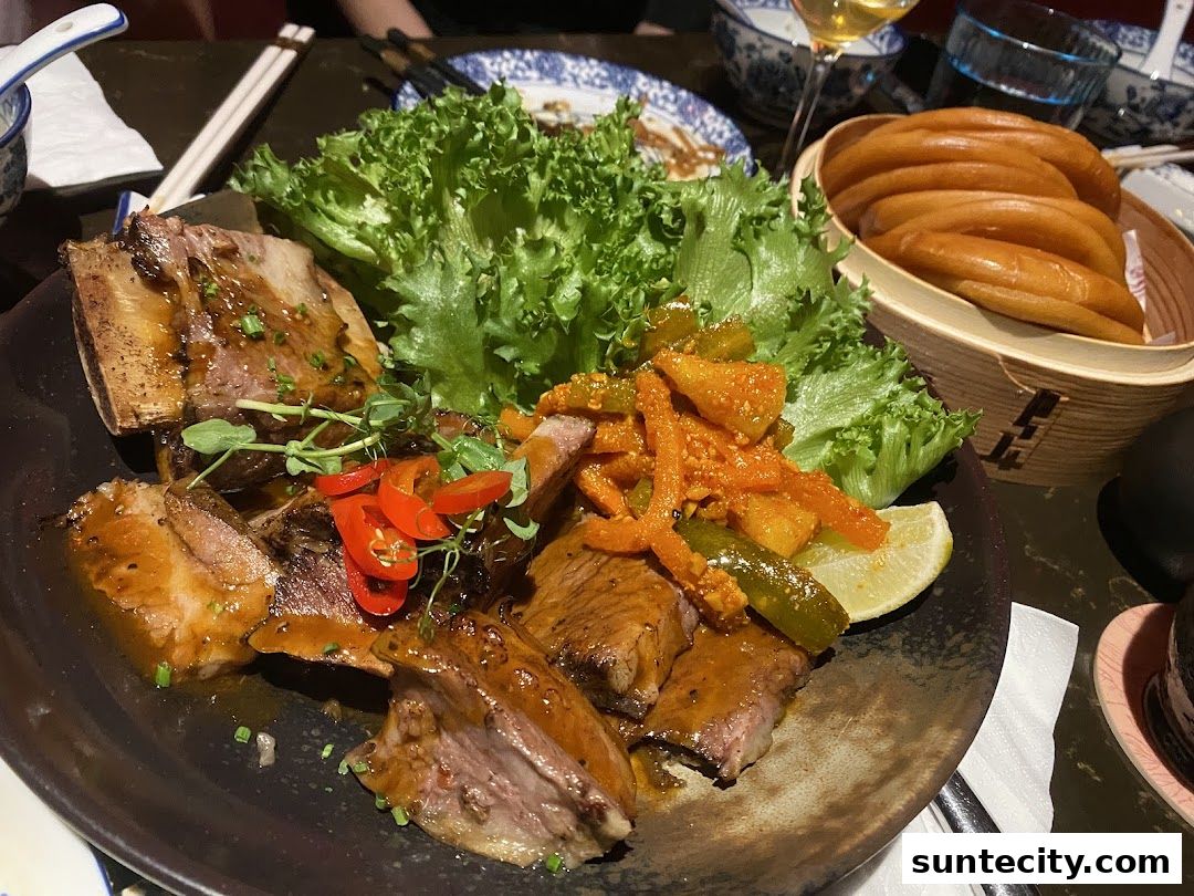 A plate of braised ribs with lettuce, pickled vegetables, and a lemon wedge, served with steamed buns.