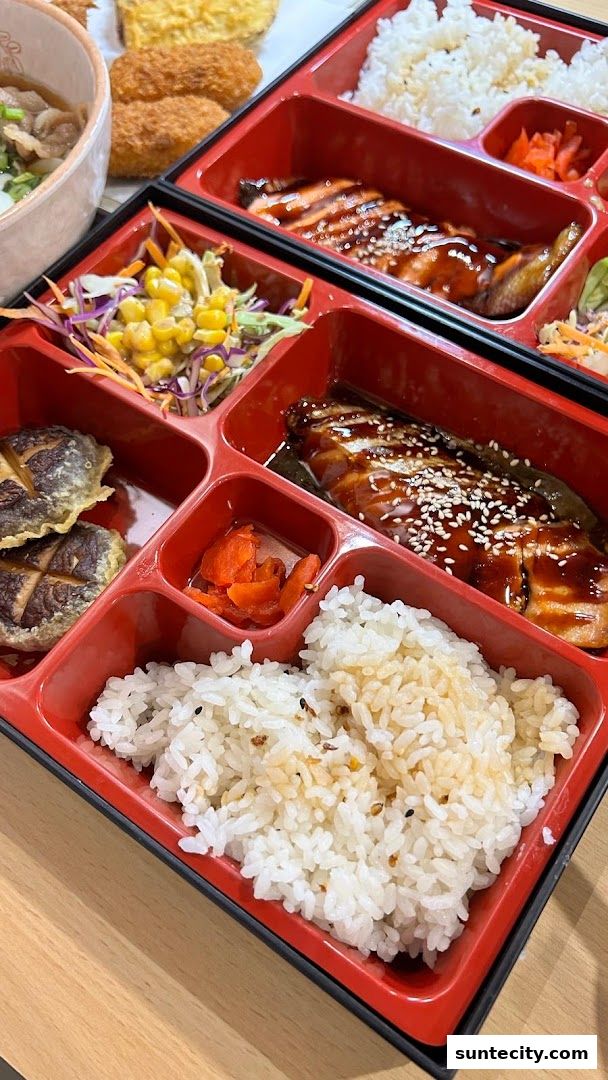 A close-up of a bento box with grilled salmon, rice, salad, and fried items.