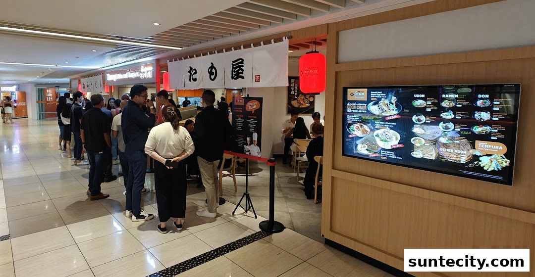 A queue of people waiting outside Tamoya Udon Tempura Suntec City, with a digital menu displayed.