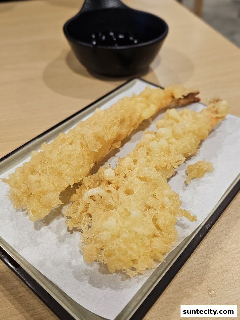 Two pieces of golden-brown tempura shrimp served on a white paper with a dipping sauce.