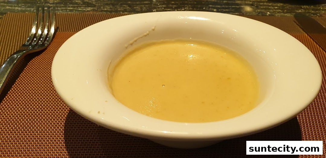 A close-up shot of a bowl of creamy yellow soup with a fork and knife on a table.