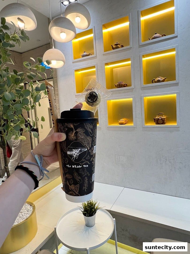 A hand holds a branded cup from The Whale Tea SG with a small pastry in front of a wall display.