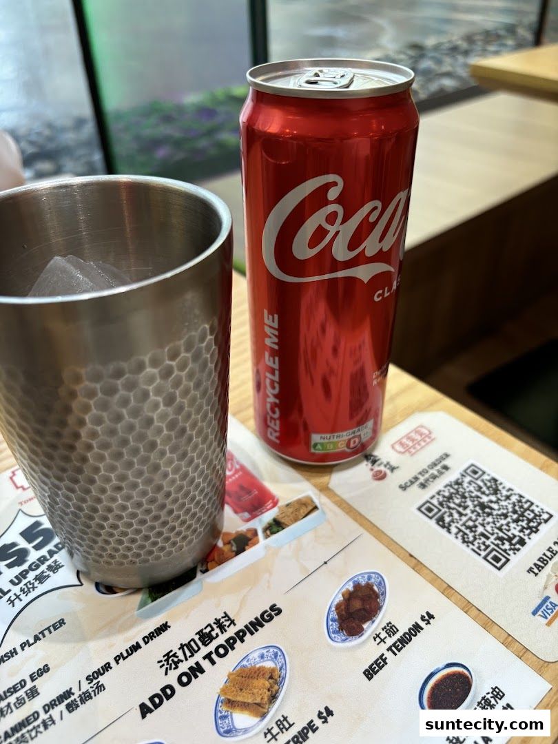 A can of Coca-Cola and a metal cup with ice sit on a table with a menu.