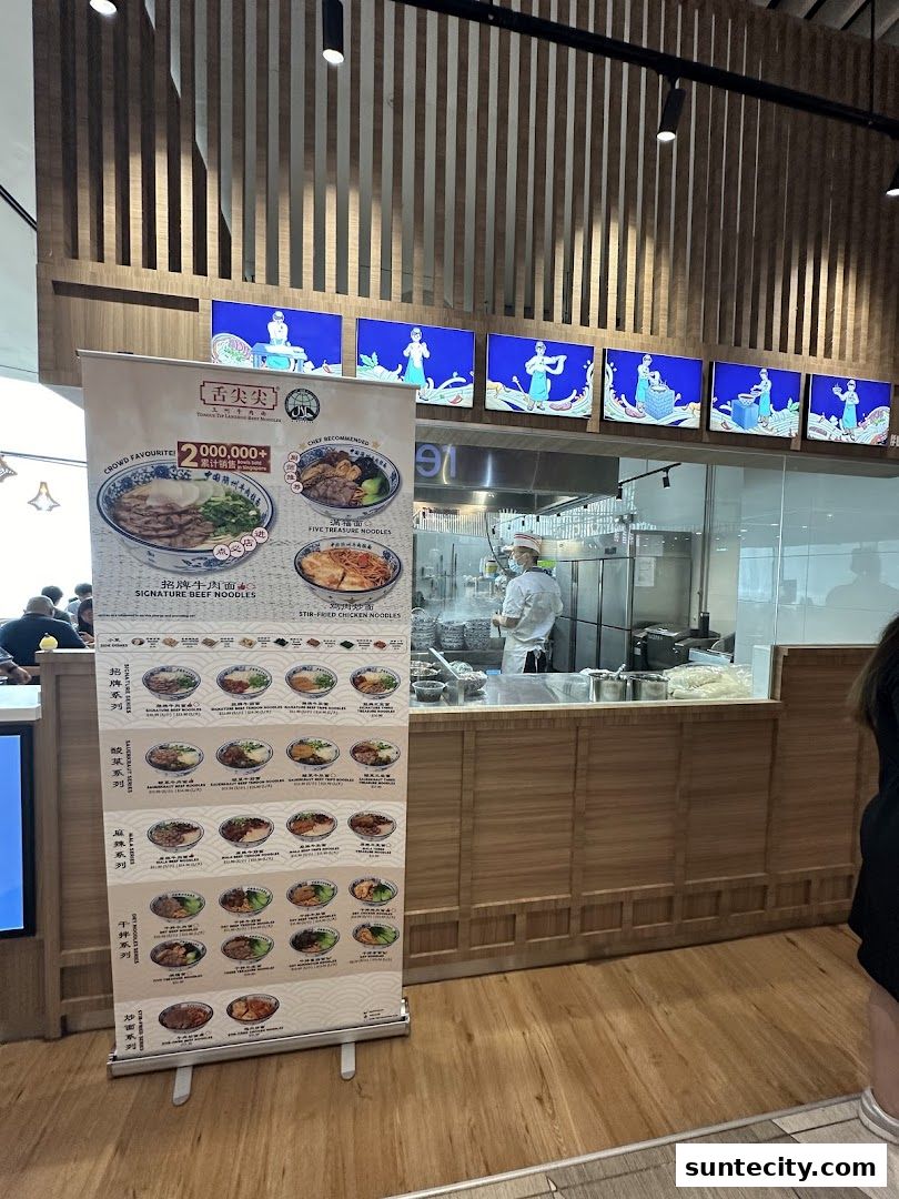 A banner displays various noodle dishes in front of a busy noodle restaurant kitchen.