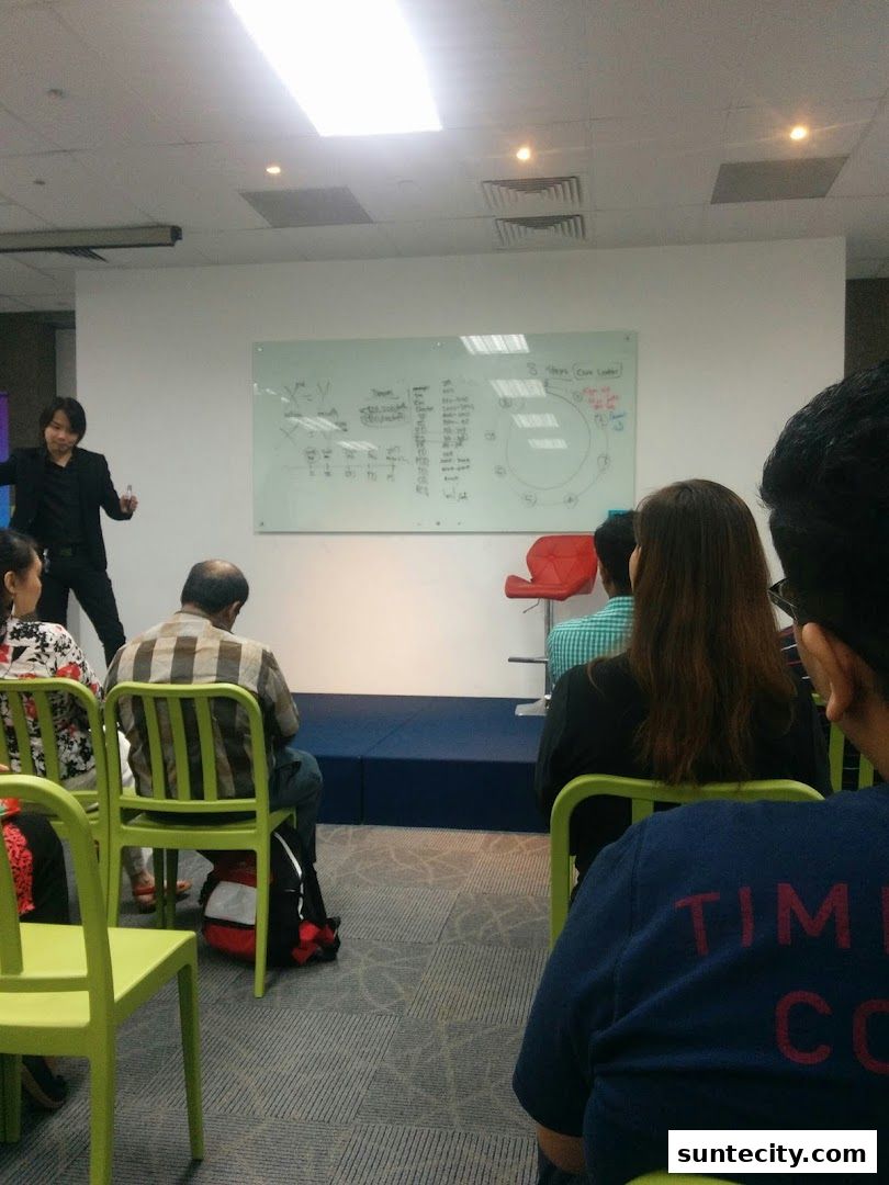 A presenter stands by a whiteboard while an audience sits in chairs.