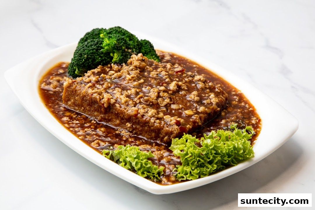 A white plate holds a dish of fried tofu topped with minced meat and sauce, garnished with broccoli and lettuce.