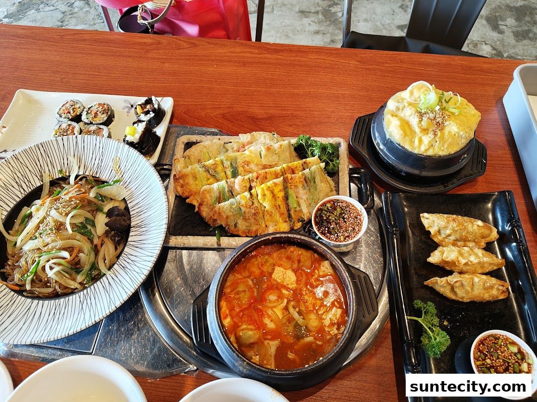 A table laden with various Korean dishes, including japchae, kimbap, pajeon, dumplings, and jjigae.