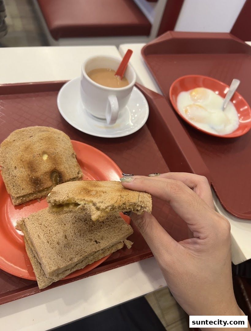 A person holding a half-eaten kaya toast sandwich with a cup of tea and soft-boiled eggs.