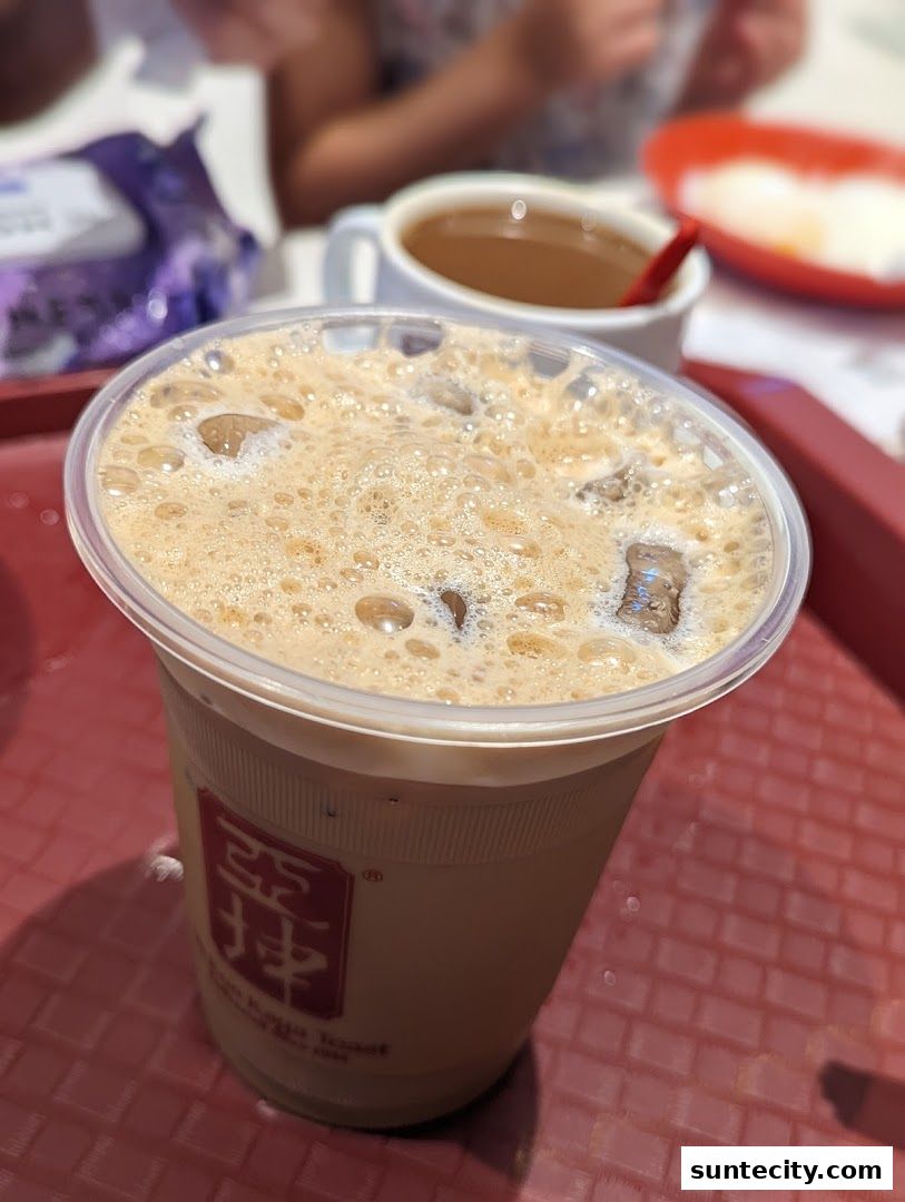 A close-up of an iced coffee with foam and ice, served in a Ya Kun Kaya Toast cup.