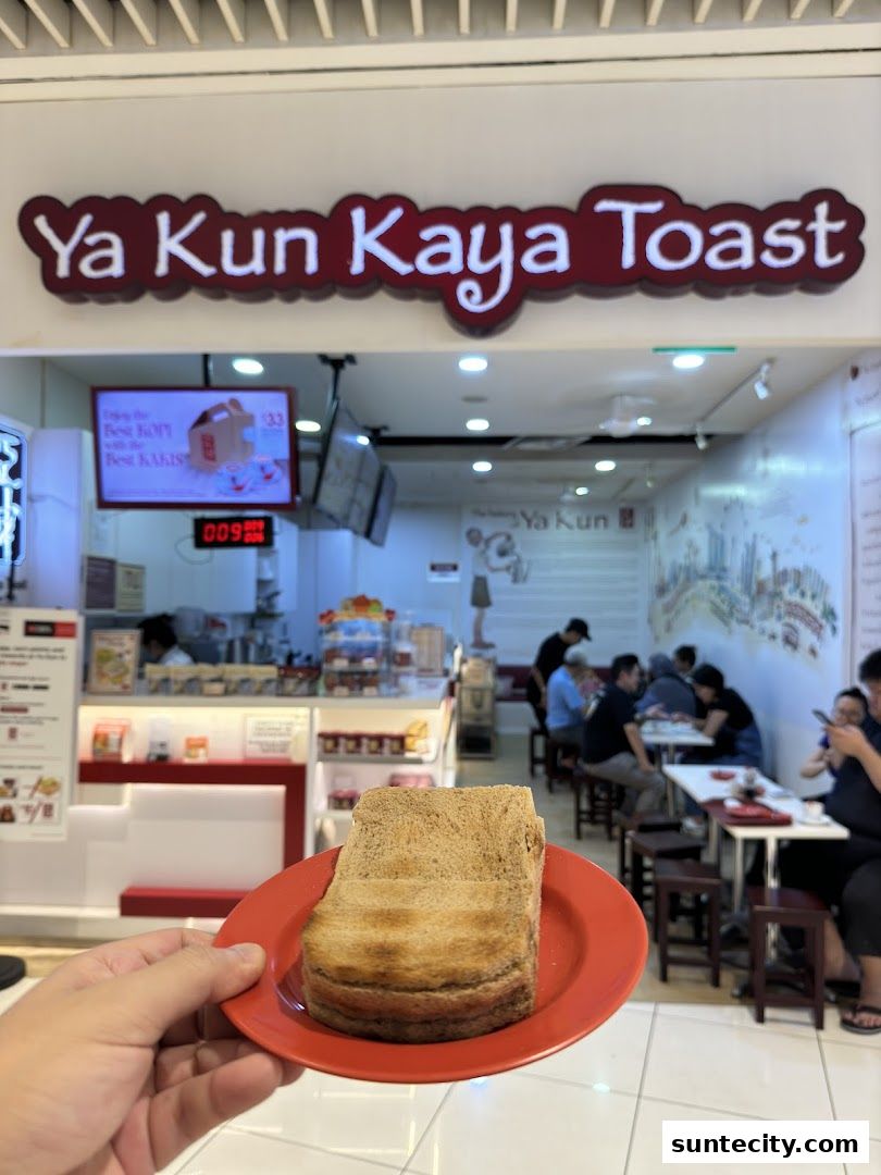 A hand holds a plate of kaya toast in front of the Ya Kun Kaya Toast shop.