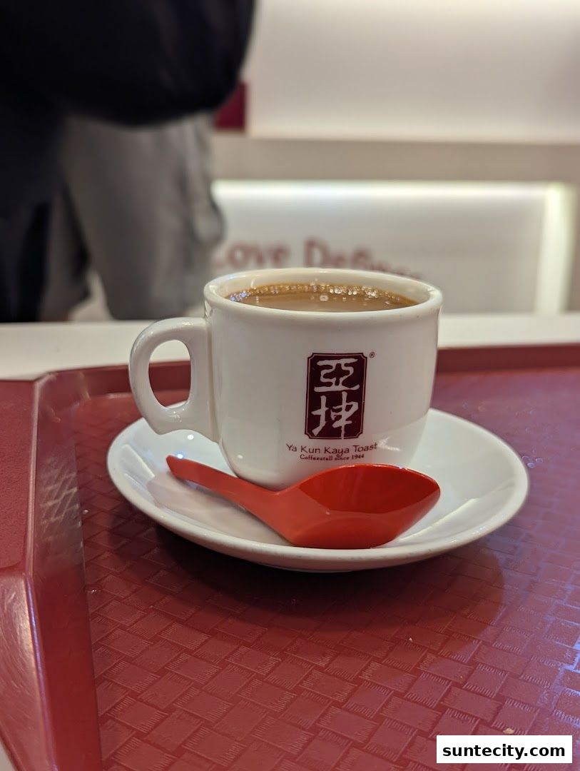 A close-up of a steaming cup of coffee with the Ya Kun Kaya Toast logo and a red spoon.